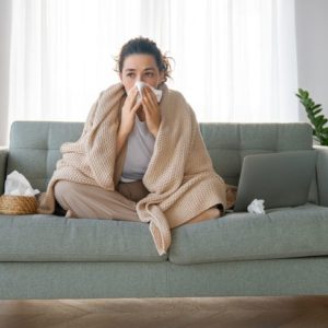 Infezioni respiratorie: come riprendersi rapidamente da influenza e raffreddore;  Una donna è seduta su un letto coperta da una coperta e lotta con i sintomi dell’influenza