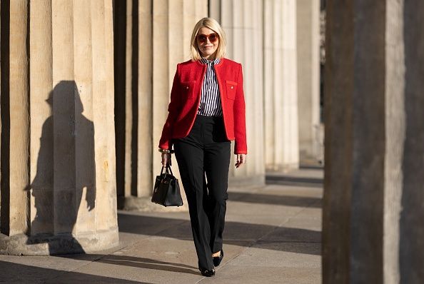 Bezvremenske dizajnerske torbe koje nikada neće izaći iz mode; Plavokosa žena u crvenom blejzeru, košulji na bruge i crnim pantalonama nosi crnu Hermes Birkin torbu