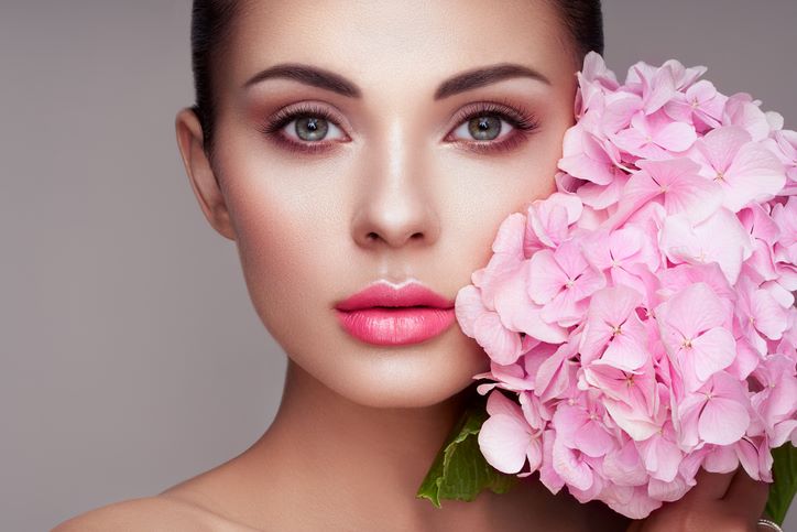 Kako odabrati savršen karmin prema tonu kože i boji kose; žensko lice u krupnom planu, perfektno našminkano, sa prelepom, ružičastom nijansom karmina i raskošnim cvetom s pink laticama sa desne strane
