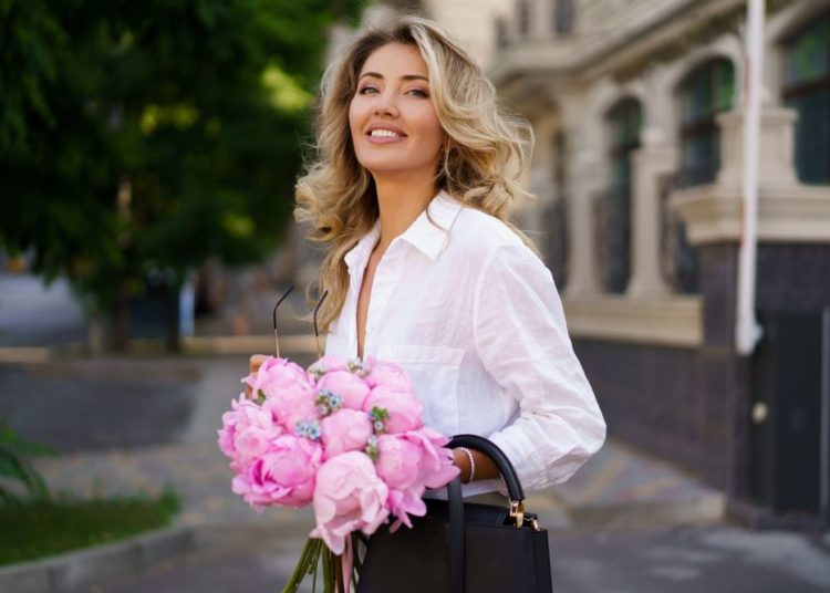 Prolećni autfiti koji izgledaju skupo a ne koštaju puno; otmena, plavokosa žena u beloj košulji i sa crnom torbom pozira sa buketom roze cveća